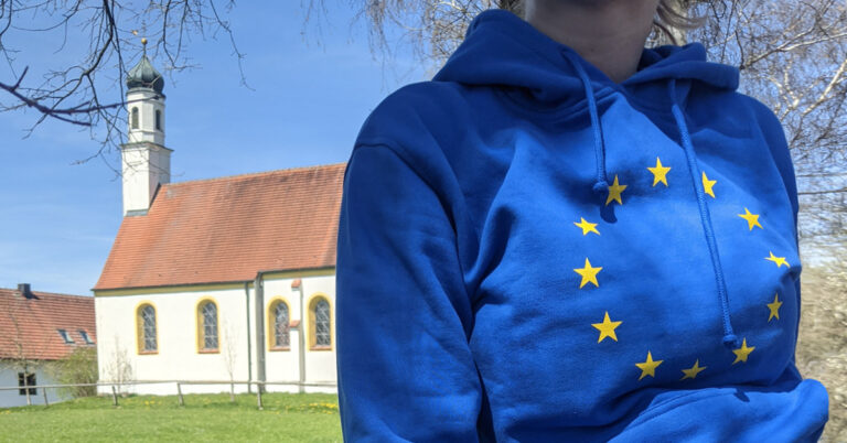 Nahaufnahme einer jungen Frau: Sie trägt einen blauen Pullover mit den Europasternen, im Hintergrund eine Kapelle mit Zwiebelturm