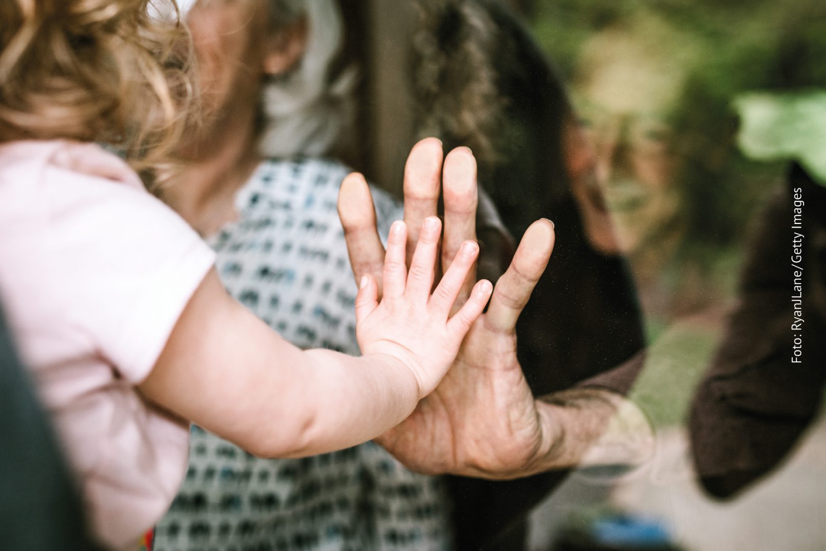 Nähe in Corona-Zeiten: Ein Kind und eine ältere Person, zwischen ihnen eine Fensterscheibe. Sie legen die Hände aufeinander.