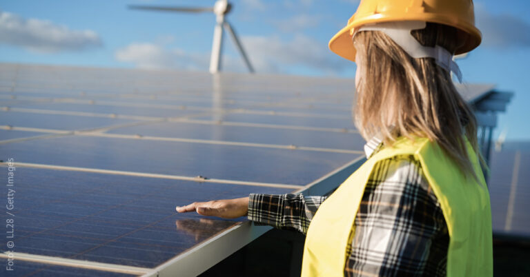 Eine Frau steht neben einer Freiflächen-Solaranlage, sie hält eine Hand über die Oberfläche, im Hintergrund steht ein Windrad