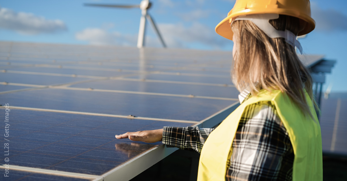 Eine Frau steht neben einer Freiflächen-Solaranlage, sie hält eine Hand über die Oberfläche, im Hintergrund steht ein Windrad