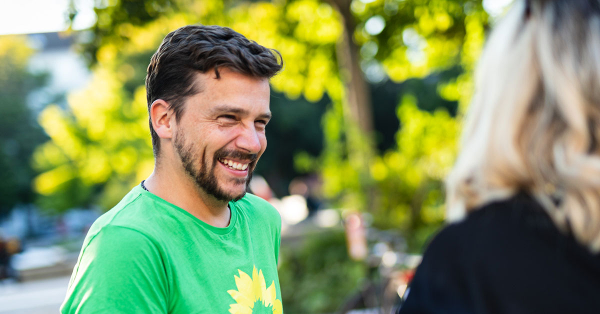 Ein Mitglied beim Flyer verteilen. Er trägt ein grünes T-Shirt mit Sonnenblume und lächelt eine Passantin freundlich an.