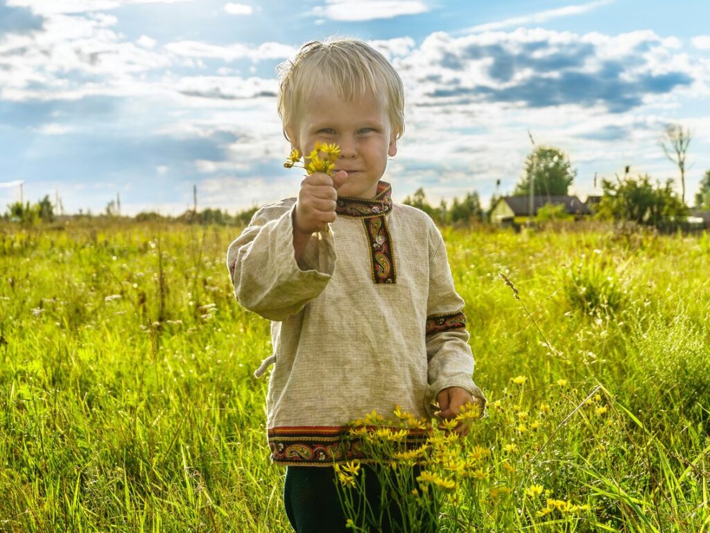 Ein kleiner, blonder Junge steht auf einer grünen Wiese und hält eine handvoll, gepflückter gelber Blumen in der Hand